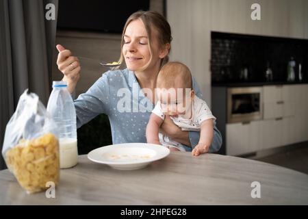 Familie beim Frühstück in einer weißen sonnigen Küche. Eine junge Mutter mit einem kleinen Sohn isst Maisflocken mit Milch, Diätkost. Elternteil mit Kleinkind Kind eati Stockfoto