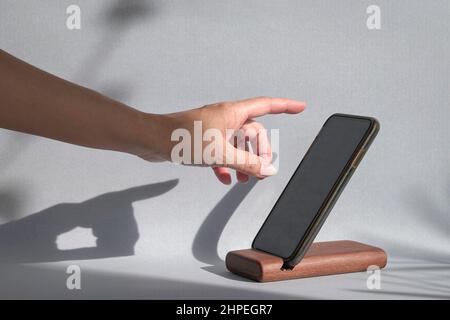 Finger, der auf ein Smartphone streut, das auf einem Halter steht, mit Schatten an der Wand. Nachbau-Remplate. Stockfoto