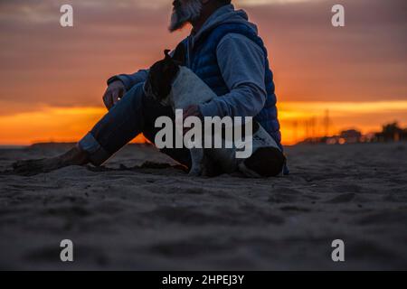 Mann mit Hund sitzt am Strand gegen den Himmel während des Sonnenuntergangs Stockfoto