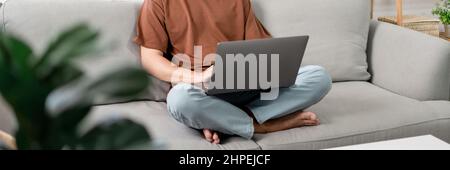 Technologiekonzept der Mann, der in legerer Kleidung auf dem Sofa sitzt, sein Notebook auf dem Schoß auslaht und seine Arbeit verrichten will. Stockfoto