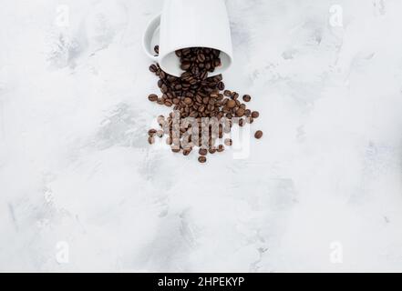 Verschüttete Kaffeebohnen aus der weißen Keramikschale auf weißem strukturiertem Hintergrund. Café-Bar, Koffein, Roastkonzept Stockfoto
