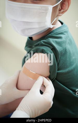 Nahaufnahme eines Arztes, der nach einer Injektion im Krankenhaus den Gips auf den Arm eines kleinen Jungen in Maske klebt Stockfoto