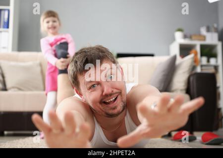 Mädchen ziehen Väter Fuß und wollen spielen, Mann schreit auf dem Teppich zu Hause Stockfoto