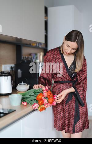 Eine glückliche Frau, die zu Hause in der Küche einen Kaffee in einem Seidengewand trinken und dabei frische Blumen genießen kann Stockfoto
