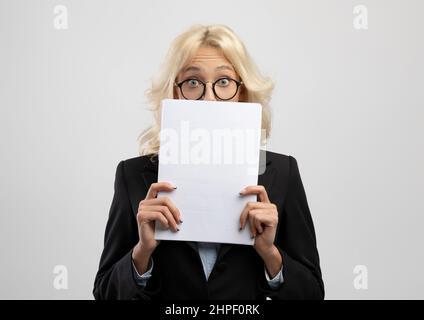 Schockierte Unternehmerin, die hinter Dokumenten guckt und sich auf hellgrauem Studiohintergrund posiert Stockfoto