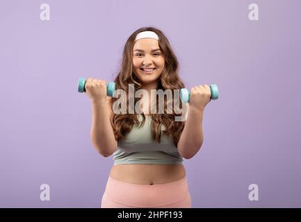 Frohes Lächeln aufgeregt plus Größe europäische Millennial Dame in Sportswear Hanteln, auf violettem Hintergrund, Studio Stockfoto