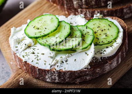 Toast mit Frischkäse, Gurke und Roggenbrot auf einem Holzbrett, dunkler Hintergrund. Stockfoto