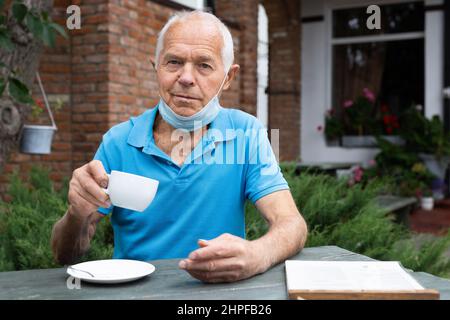 Älterer Mann in Schutzmaske mit einer Tasse Kaffee oder Tee in der Hand sitzt am Tisch des Straßencafés und liest die Speisekarte Stockfoto