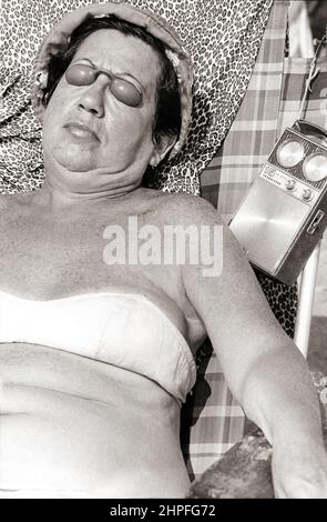 Eine Frau, die in Brighton Beach sonnenbaden und dabei einem Transistor-Radio lauschen und Augenschutz aus Kunststoff tragen. In Brooklyn, New York, um 1976. Stockfoto