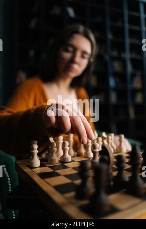 Vertikale selektive Fokusaufnahme einer jungen Frau mit eleganten Brillen, die Schach bewegen lässt, die auf einem Sessel im dunklen Raum sitzt. Stockfoto