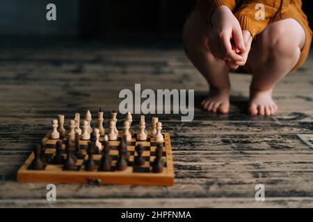 Die Beine einer nicht erkennbaren Frau, die in der Nähe des Schachbretts auf dem Holzboden im dunklen Raum sitzt und an Schachzug denkt, sind mit einem abgeschnittenen Schuss übersät. Stockfoto