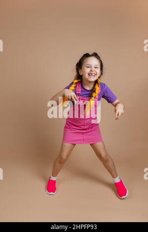 Entzückend glückselig glänzendes weibliches Kind mit Kanekalon Zöpfen springen Spaß lächeln mit fehlenden Zahn gekleidet in rosa Jumpsuit, lila T-Shirt Stockfoto