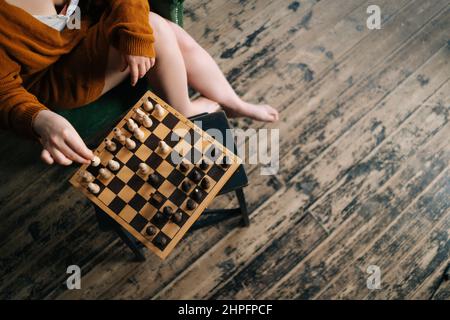 Nahaufnahme von oben, in der sich eine nicht erkennbare Frau auf einem Sessel in einem dunklen Raum mit Vintage-Holzboden befindet, die Schach bewegt. Stockfoto