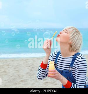Porträt der schönen jungen hübschen, fröhlichen, niedlichen, blonden Frau im gestreiften Hemd, Denim-Overalls mit Seifenblasen am Meer Stockfoto