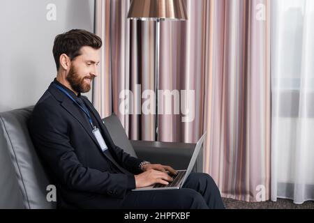 Fröhlicher Geschäftsmann mit ausweis, der seinen Laptop benutzt, während er auf der Ledercouch im Hotelzimmer sitzt Stockfoto