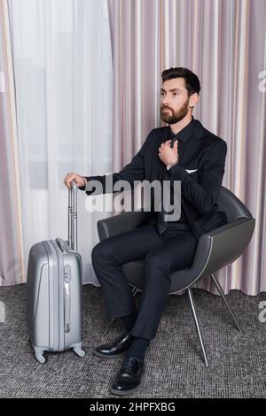 Bärtiger Geschäftsmann im Anzug, der die Krawatte anpasst und auf einem Sessel neben dem Gepäck im Hotelzimmer sitzt Stockfoto