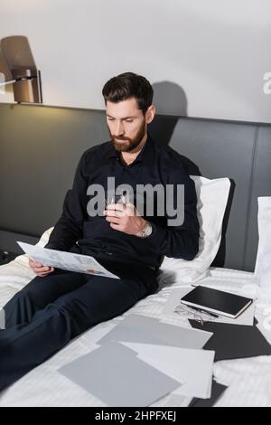 Bärtiger Mann mit einem Glas Whiskey und sitzt im Hotelbett in der Nähe von Infografiken Stockfoto