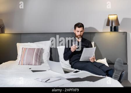 Bärtiger Mann, der ein Glas Whiskey in der Hand hielt und sich das Dokument in der Nähe von Gadgets auf dem Hotelbett ansah Stockfoto