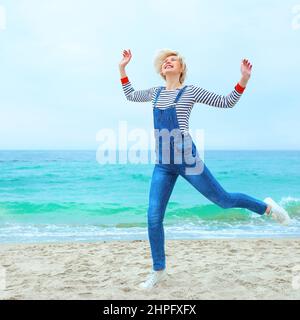 Schöne junge blonde kaukasische Frau im Urlaub in gestreifter Bluse, Sneakers und Denim insgesamt am Strand durch die erstaunliche blaue Meer Hintergrund. Stockfoto