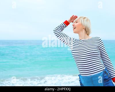 Schöne junge blonde kaukasische Frau im Urlaub in gestreifter Bluse, Sneakers und Denim insgesamt am Strand durch die erstaunliche blaue Meer Hintergrund. Stockfoto