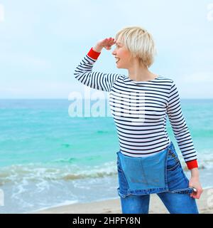 Schöne junge blonde kaukasische Frau im Urlaub in gestreifter Bluse, Sneakers und Denim insgesamt am Strand durch die erstaunliche blaue Meer Hintergrund. Stockfoto