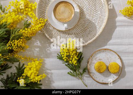 Zweige gelber Mimosenblüten auf weißen Leinenbettlaken, Tasse oder Kaffee, französische gelbe Makronen. Gruß zum Internationalen Frauentag am 8t. März Stockfoto