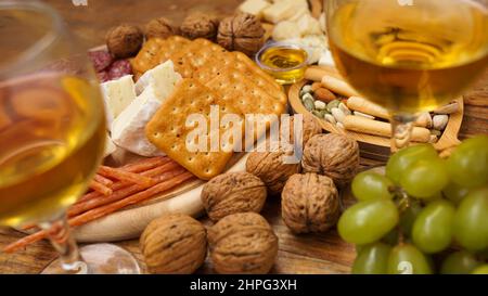 Snacks für Wein. Käse- und Fleischplatte. Wurst, Käse, Nüsse, Trauben, Cracker auf Holzgrund. Zwei Gläser Weißwein. Stockfoto