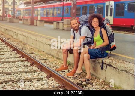 Glückliche junge und schöne Touristen mit Rucksäcken sitzen am Bahnhof in der Nähe der Gleise und genießen ihre Reise Stockfoto