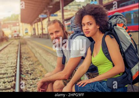 Glückliche junge und schöne Touristen mit Rucksäcken sitzen am Bahnhof in der Nähe der Gleise und genießen ihre Reise Stockfoto