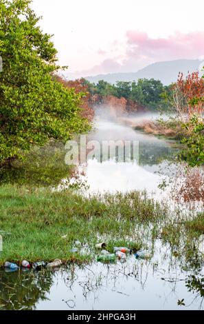 Verschmutzung von Plastikflaschen und anderen Trümmern in einem Fluss mit Nebel am Horizont. Vermüllung der Umwelt, die zu mehr Überschwemmungen führt Stockfoto
