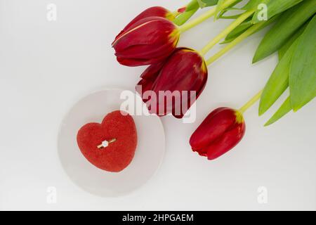 Ein Bouquet aus roten Tulpen neben einem Teller mit einem herzförmigen Kuchen, in dem ein Goldring mit einem Diamanten. Stockfoto