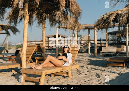 Eine junge kaukasische Frau im Badeanzug ruht im Sommer auf einer Sonnenliege am Sandstrand auf einer Sonnenliege unter einem Strohschirm. Platz kopieren. Stockfoto