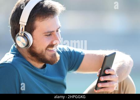 Fröhlicher Mann mit Kopfhörern, der Musik hört und im Freien auf dem Smartphone nachhört Stockfoto