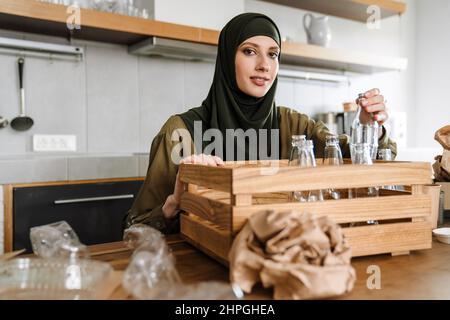 Lächelnde islamische Frau in Hijab sortiert Glasflaschen und Plastikbehälter für das Recycling in der Küche Stockfoto