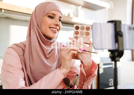 Junge muslimische Frau in Hijab vloggt sich über Beauty-Produkte mit einem Video für ihren Blog über Kosmetik mit Handy-Kamera zu Hause zu machen Stockfoto