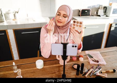 Junge muslimische Frau in Hijab vloggt sich über Beauty-Produkte mit einem Video für ihren Blog über Kosmetik mit Handy-Kamera zu Hause zu machen Stockfoto