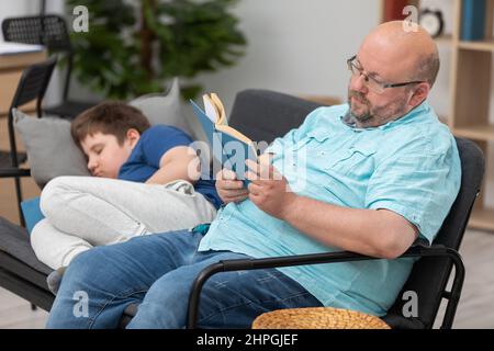 Papa liest ein Buch und Sohn schläft beim Lesen ein. Stockfoto