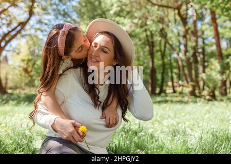 Mädchen küsst Mutter auf Wangenumarmungen, während sie im Frühlingspark auf Gras sitzt und Blumen schenkt. Familie geht im Freien spazieren. Muttertag Stockfoto