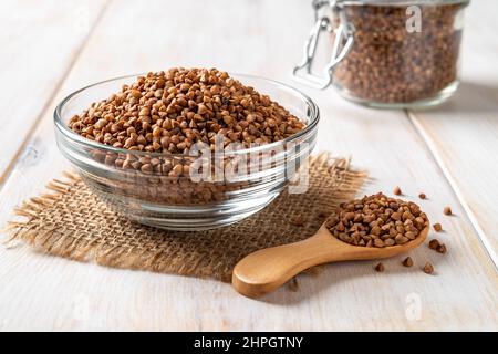 Zutat zum Kochen Buchweizenbrei Konzept. Roher Bio-Buchweizen in einer Glasschale und Holzlöffel auf einer weißen Holzplatte. Glutenfreie Ernährung Stockfoto