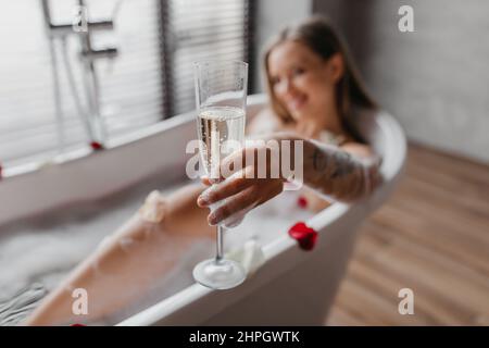 Junge Frau hält ein Glas Champagner, während sie sich zu Hause im Schaumbad ausruhen, und konzentriert sich auf das Getränk Stockfoto