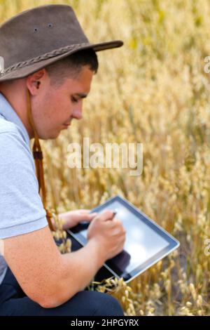 Digitale Farm. Porträt eines Bauern, der im goldenen Weizenfeld sitzt und auf dem Tablet scrollt. Junger Mann mit Cowboyhut auf dem Feld, der Weizenernte untersucht. Hafer Stockfoto