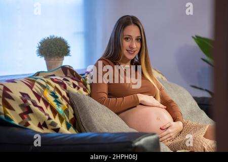 Schwanger junge Frau sitzt auf dem Sofa berühren ihren Bauch und Blick auf die Kamera Stockfoto