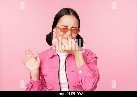 Stinkend. Asiatisches Mädchen in Sonnenbrille und stilvollem Outfit, schließt ihre Nase vor Ekel, mag schlechten Geruch nicht und steht über rosa Hintergrund Stockfoto