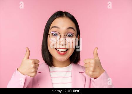 Super, Glückwunsch. Gesicht einer aufgeregten asiatischen Geschäftsfrau in einer Brille, lächelnd zufrieden, mit Daumen nach oben, stehend über rosa Hintergrund Stockfoto