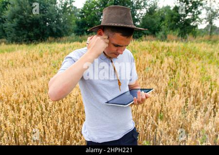 Junger Bauer. Porträt eines Bauern, der im goldenen Weizenfeld sitzt und auf dem Tablet scrollt. Junger Mann mit Cowboyhut auf dem Feld, der Weizenernte untersucht. Hafer Stockfoto