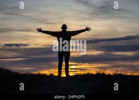 Silhouette eines Mannes, der mit ausgestreckten Händen gegen den romantischen Sonnenuntergangshimmel steht, Konzept von Freude und Glück Stockfoto