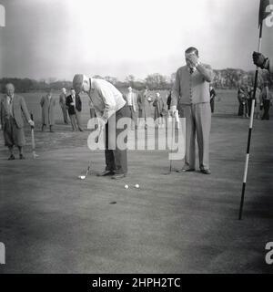 1950s, historisch, spielt an einem Firmengolftag, zwei männliche Golfer auf einem Puttin-Grün, einer im Begriff, seinen Ball in Richtung des Lochs zu setzen, Port Talbot, Wales, Großbritannien. Stockfoto