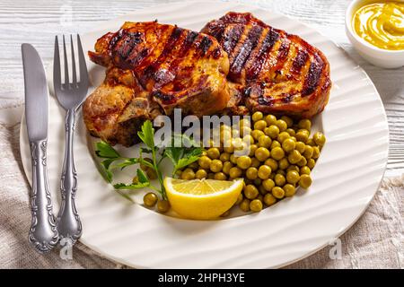 Nahaufnahme von gegrillten Schweinefleisch-T-Bone-Steaks mit gekochten grünen Erbsen und Zitrone auf einem weißen Teller auf einem Holztisch mit Senf, Stockfoto