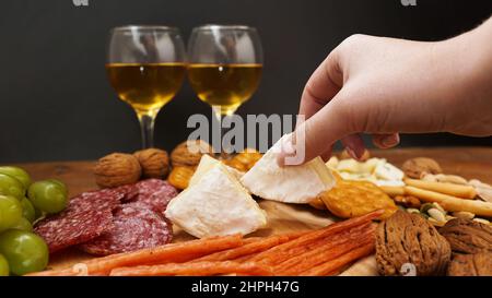 Eine Frau nimmt ein Stück Camembert-Käse von einem Käseplatte. Italienische Käse- und Fleischplatte für einen Weinsnack. Trauben, Nüsse, Cracker. Stockfoto