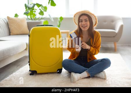 Aufgeregte junge Dame, die mit dem Koffer auf dem Boden sitzt, ihren Pass und das Flugticket vorzeigt und sich auf Reisen ins Ausland vorbereitet Stockfoto